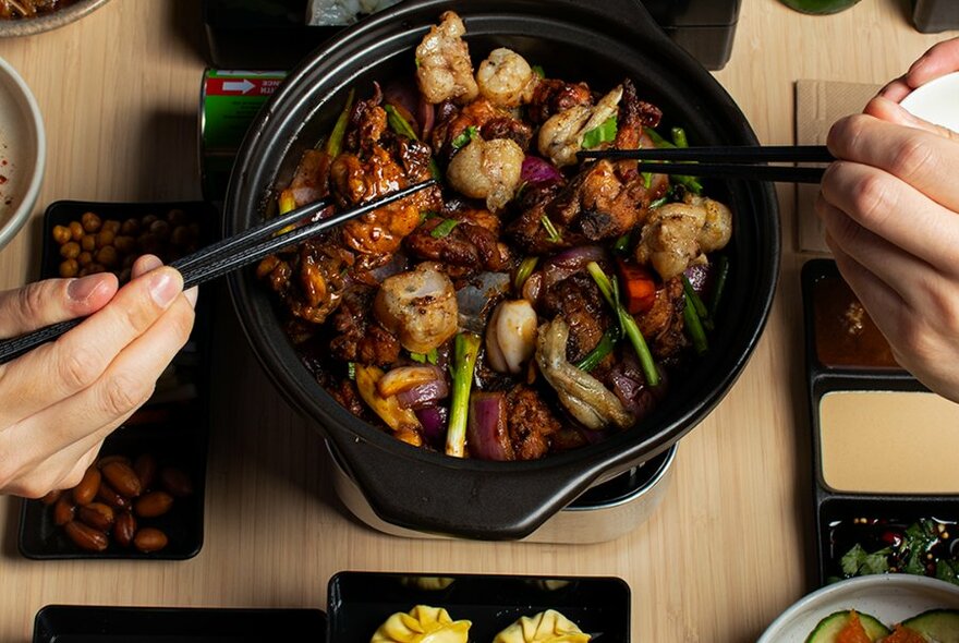 Claypot of chicken and vegetables cooking on a burner on a dining table, with two hands selecting food out of the claypot using chopsticks.