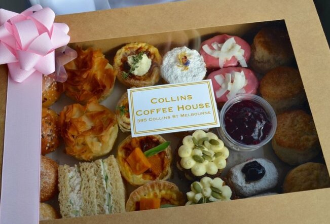 A box with a pink ribbon and bow on the top left corner. Inside the box are assorted pastries, cakes, scones and sandwiches. A sticker on the front says 'Collins Coffee House'.