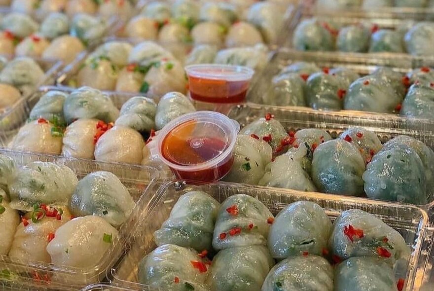 Takeaway chilli sauce containers balanced on top of trays of spinach and prawn dumplings in plastic takeaway containers.