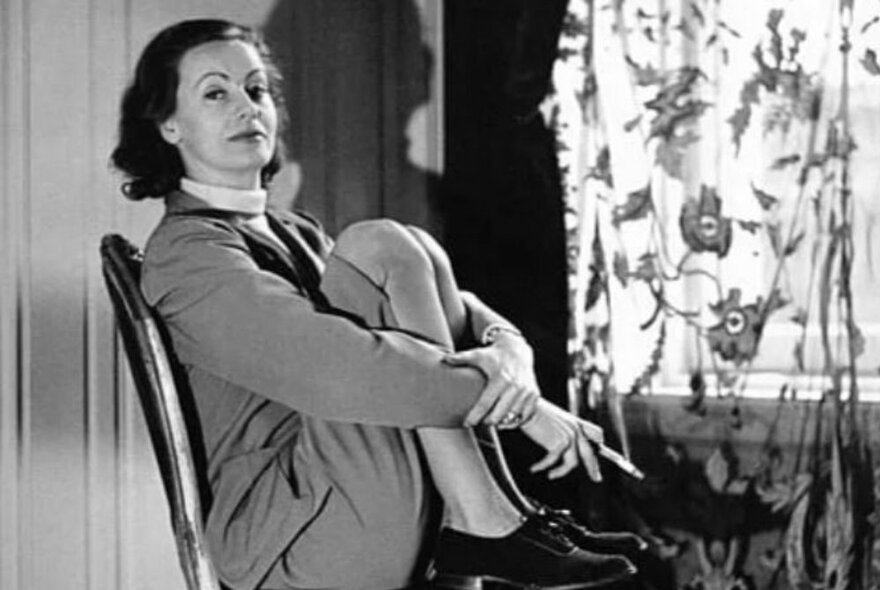 Fashion photograph of Greta Garbo seated on a chair with her knees drawn up to her chest, wearing a suit and lace-up shoes, smoking a cigarette.