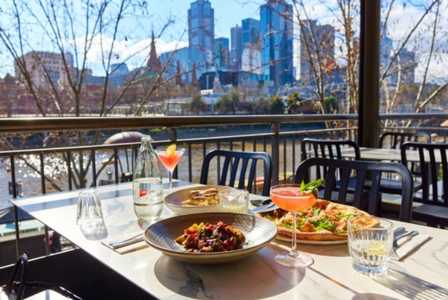 Table with pizza, bowl of pasta and cocktails seated outside by the Yarra River and city skyline background