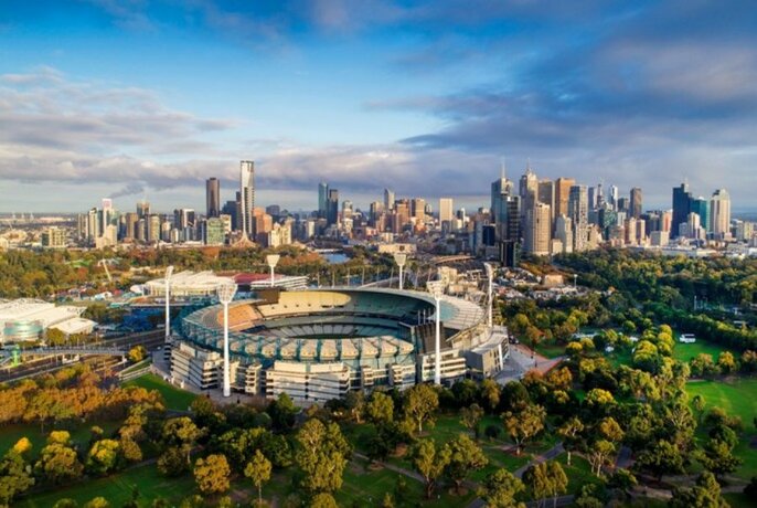 Aerial view of the MCG, surrounded by green parklands, with the Melbourne city skyline in the background.