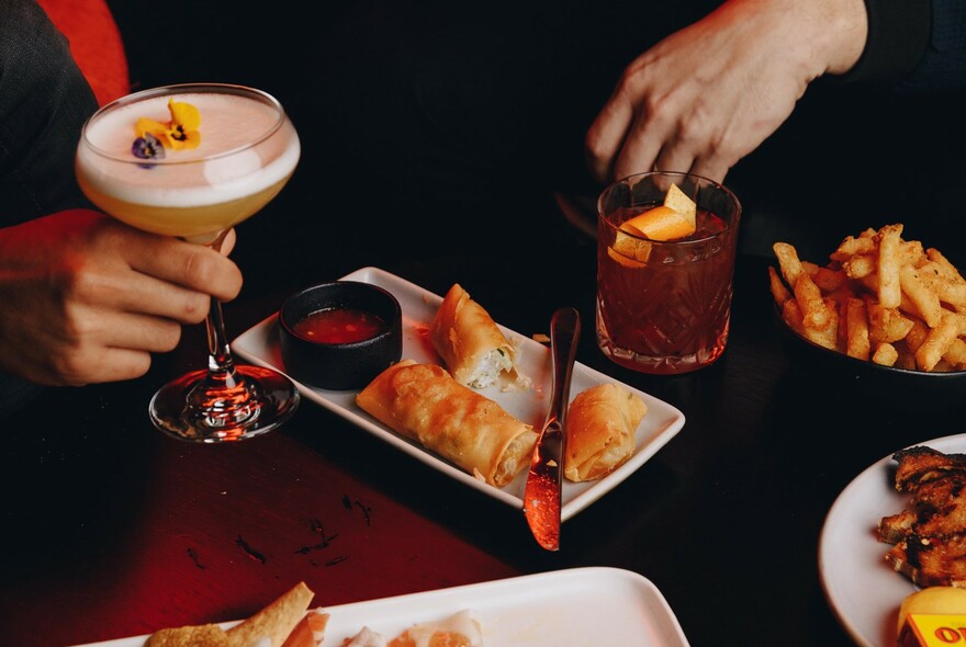 One hand holding a whisky sour cocktail, another using chopsticks to select items from plates of chips and other snacks.