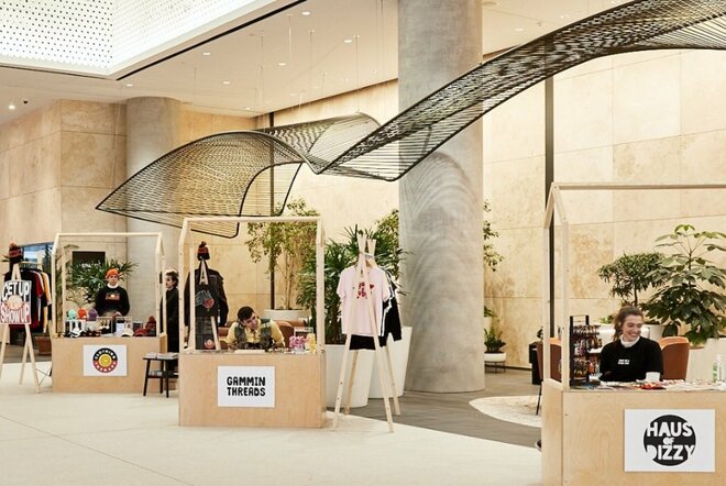 Interior airy commercial building lobby with several market stalls arranged in the space, selling clothing, jewellery and other items; people seated behind the stalls smiling.