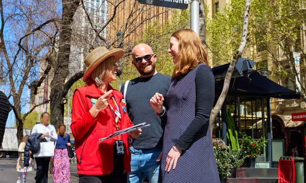 Two people standing on a sunny city street, talking to a tourism volunteer who is wearing a red jacket
