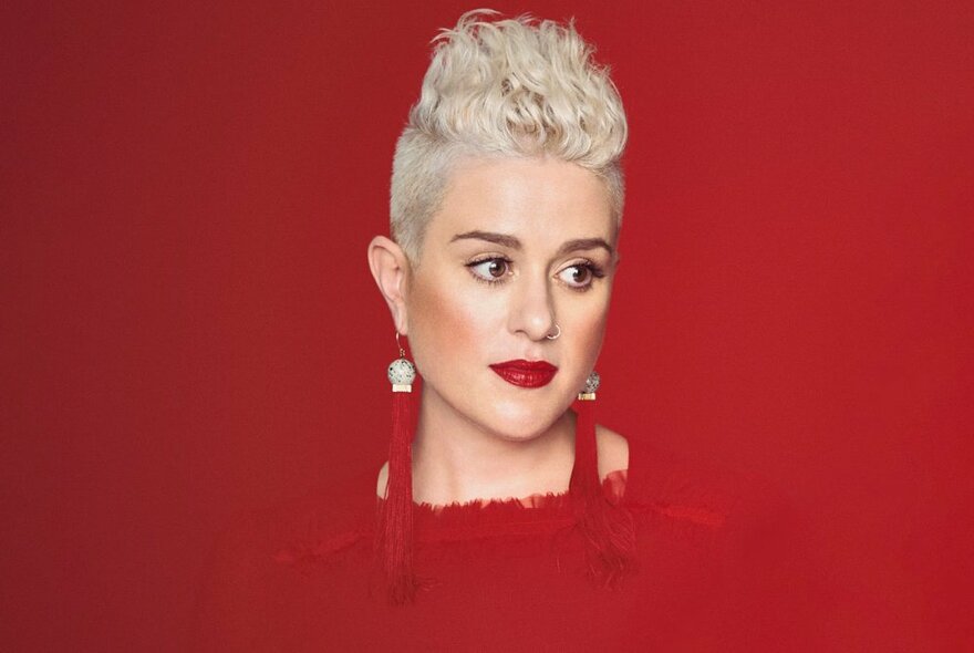 Singer Katie Noonan disappearing into a red background, wearing a matching red dress, earrings and lipstick, with blonde mohican-style hair.