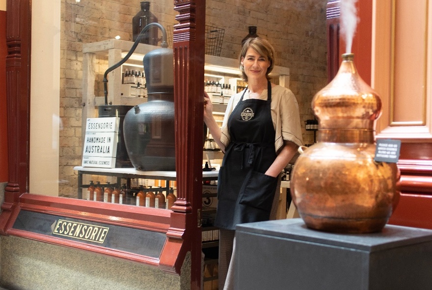A woman wearing an apron is standing in a shop doorway, in front of her is a copper urn expelling mist.