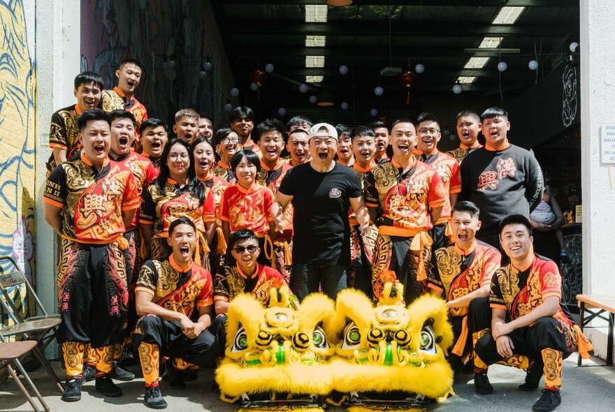 A group of people in matching black and red costumes posing with a large lion dance puppet head, at a warehouse door entrance.