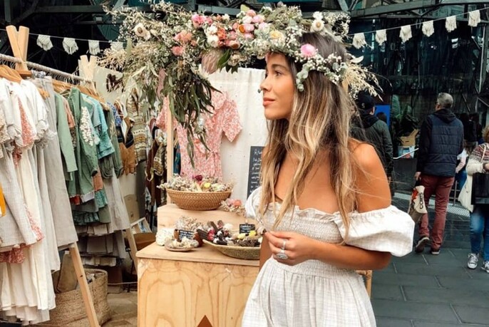 Bohemian woman with off-the-shoulder shirred gown, long blonde hair and floral headband, standing in a market with racks of gowns and ornaments.