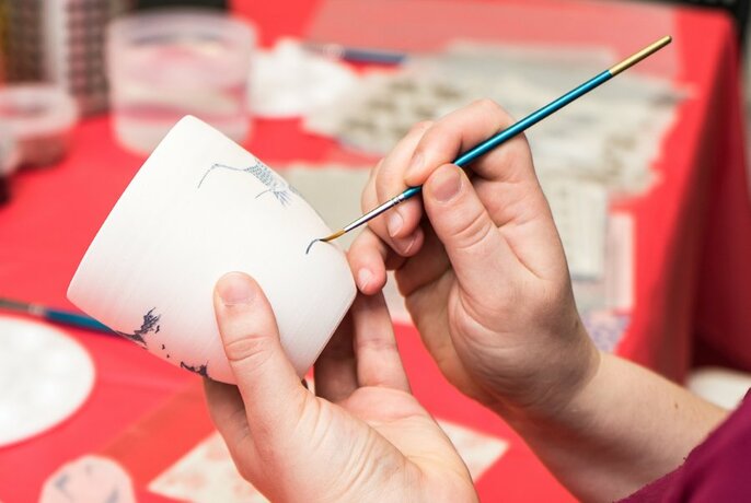 Hand holding a fine brush and decorating a small white ceramic cup with paint.