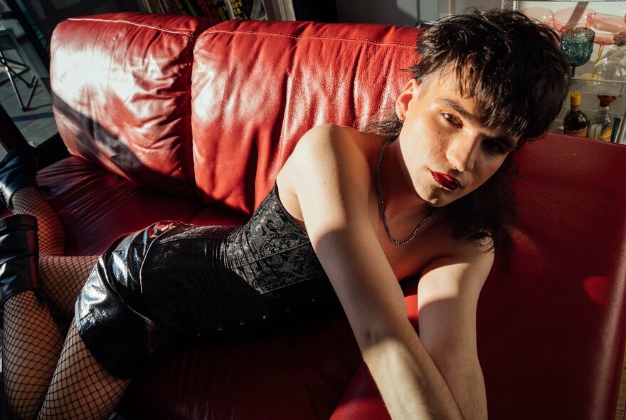 An actor posing on a red leather couch, wearing a black minidress and fishnet stockings.