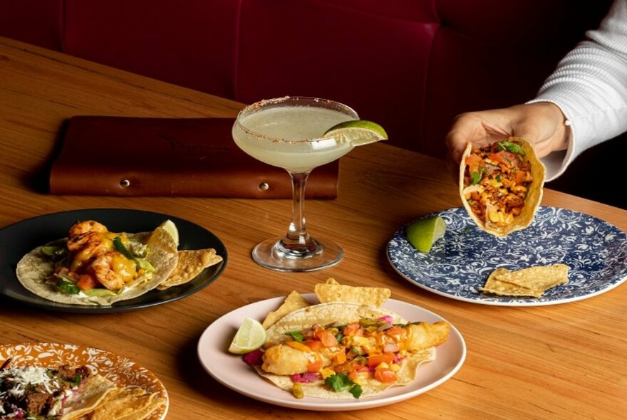 A hand picking up a filled taco from a ceramic plate on a table, with other plates of food on the table, alongside a glass of margarita garnished with a wedge of lime.