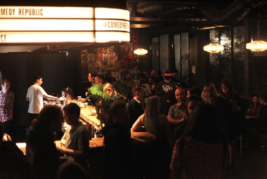 Overhead view of the bar at Comedy Republic with patrons standing around drinking and socialising, and a bartender pouring drinks under the well-lit bar area.