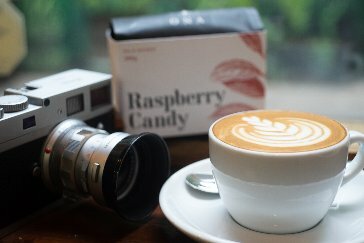 A close up shot of a coffee with latte art, a camera and a box of raspberry candy