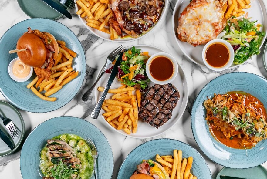 Overhead view of a table filled with plates of food including a burger and fries, a steak, fries and salad, a plate of pasta, a chicken parmigiana and more.