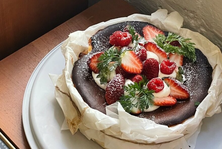 Backed cheesecake covered in cream and strawberries, wrapped in baking paper and string on plate, seen from above.