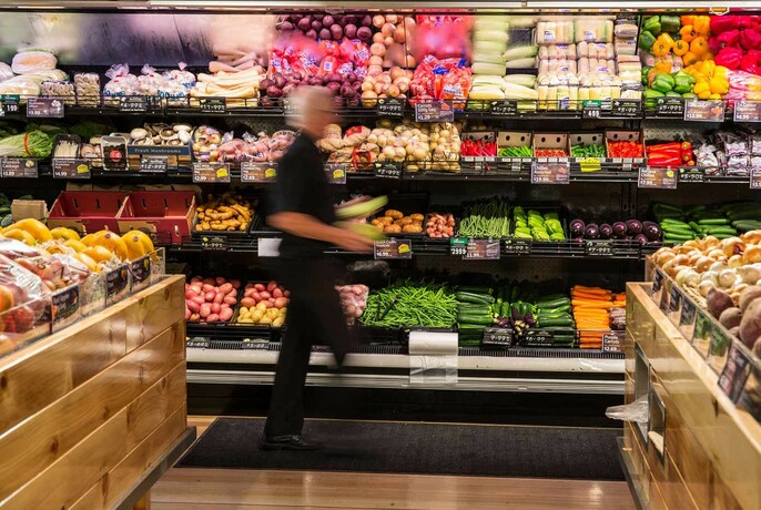 Person walking past rows of fresh produce inside a supermarket.