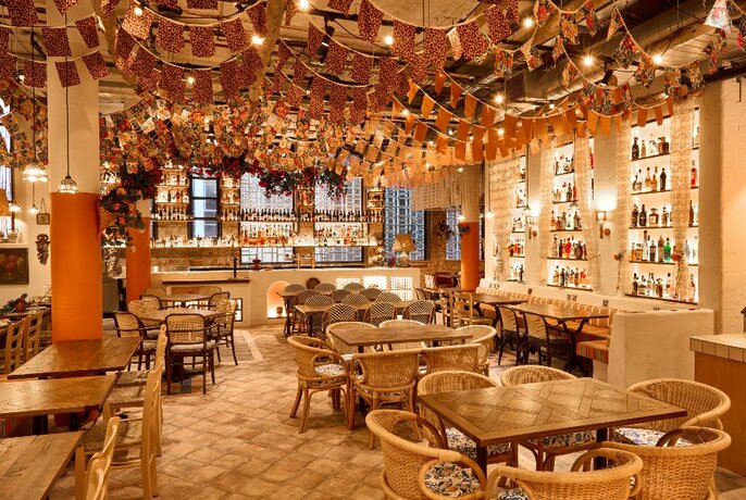 Interior of restaurant space with festive ceiling bunting, walls filled with glass bottles and large tables and cane rattan chairs.