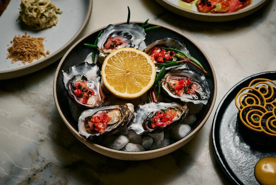 Six oysters in a bowl around half citrus fruit; other dishes partly visible.