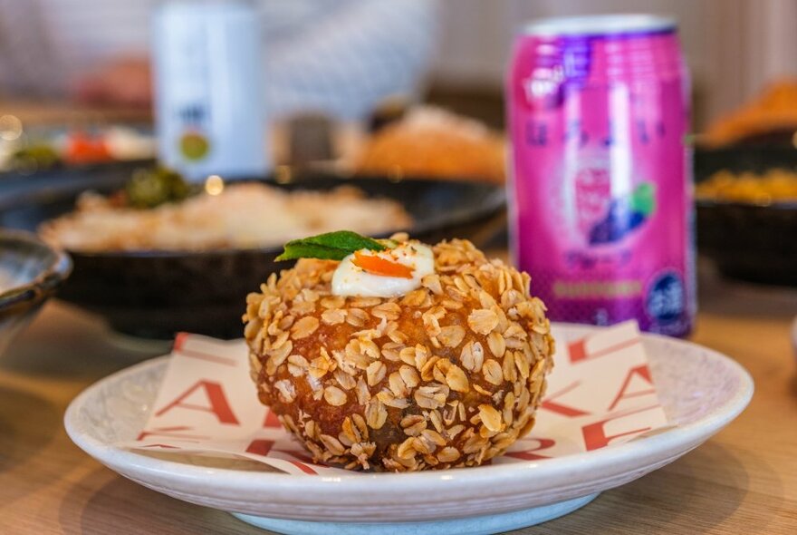 A whole round curry pan ball, resting on a plate, a pink coloured can of drink in the background, on a table in a cafe.