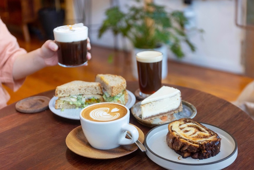 Table of coffees, sandwich and cakes, one hand holding coffee raised over table to left.