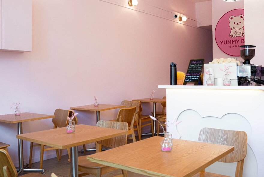 Inside the Yummy  Bear Bakery, with wooden tables and chairs, a white servery and the pink Yummy Bear Bakery sign in the background.