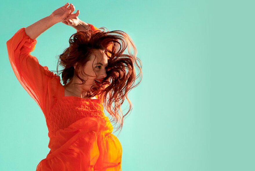 Actor wearing a bright orange top, long hair freely flowing, jumping up with her arms raised above her head, against a mint green backdrop.