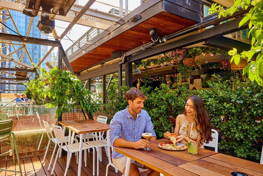 A couple sharing pizza and cocktails on a leafy rooftop bar.