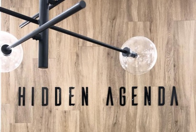 Modern ceiling lamp above Hidden Agenda logo on wall.