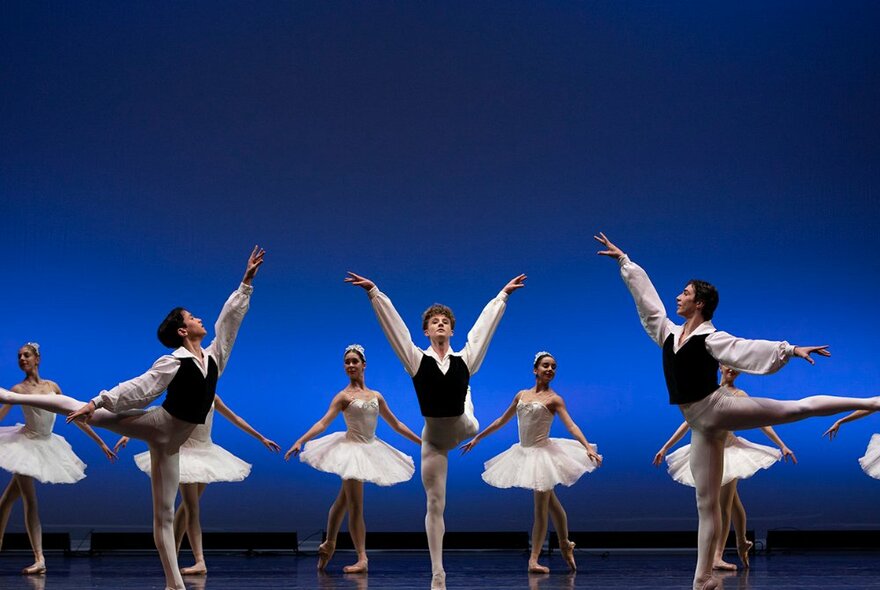 Three male ballet dancers on stage in arabesque, with five female dancers in white tutus behind.