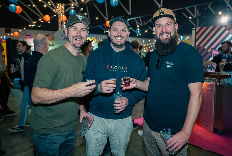 Three men in caps all trying a drink in an indoor venue with coloured lanterns.