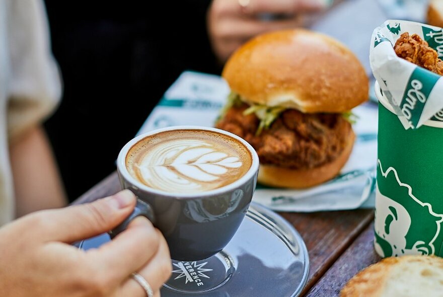 A hand holding a cup of coffee, next to the coffee is a burger and other food resting on a table top.