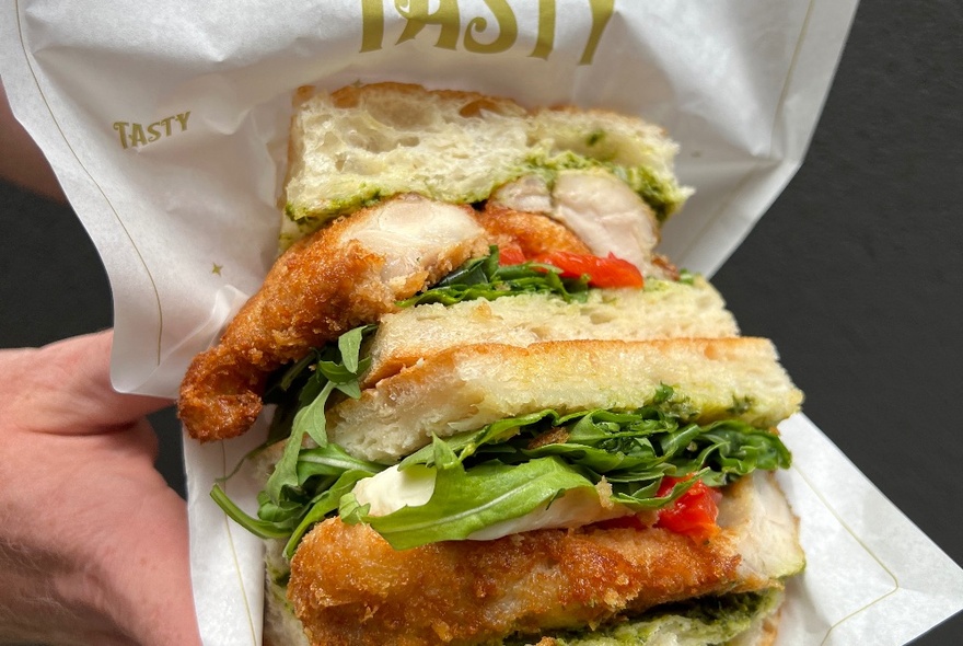 A schnitzel sandwich cut in half and being held in wrapping paper, hands just visible under the paper.