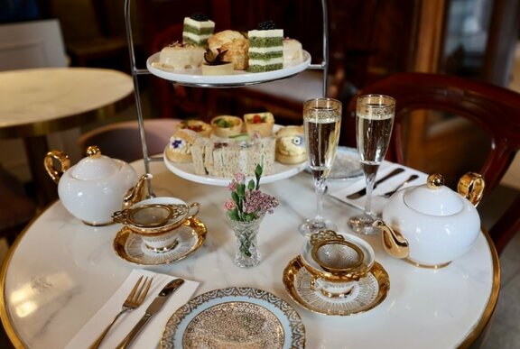 A table laid with white linen, and a high tea service with crockery, a three-tiered plate of sweet and savoury treats and two glasses of sparking wine.