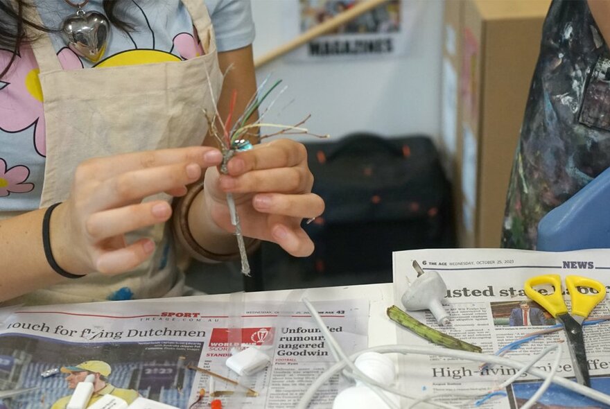 Children's hands making art at a table, with scissors, sticks and other arts and craft objects resting on a newspaper covered tabletop.