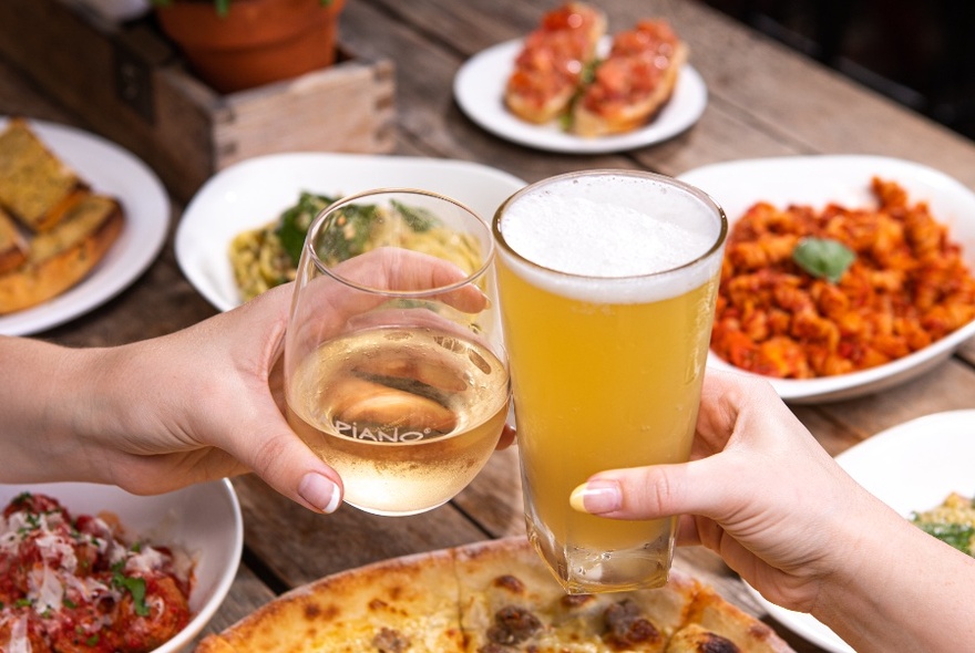 Two hands clinking together a glass of beer and a glass of wine over a table of food that includes pizza and pasta.