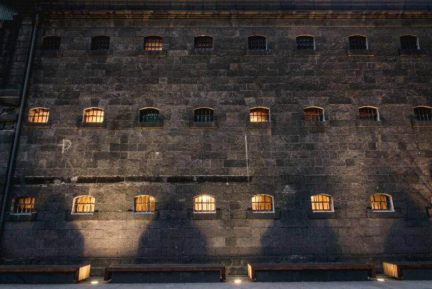 Shadows of figures on the jail's bluestone wall, with the barred windows of cells lit from within.