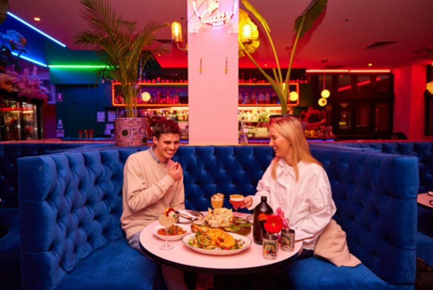 A couple sitting in a blue velvet booth laughing under neon lights, sitting around a white round table full of food and drinks.