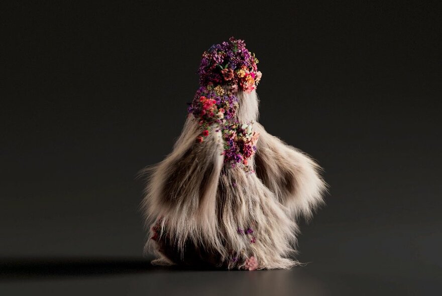 A completely fur covered creature, flowers at their head, walking past a dark wall.