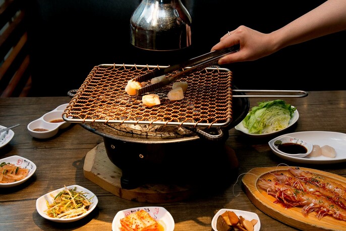Hands using tongs to add food to a grill cooking over a hot pot.