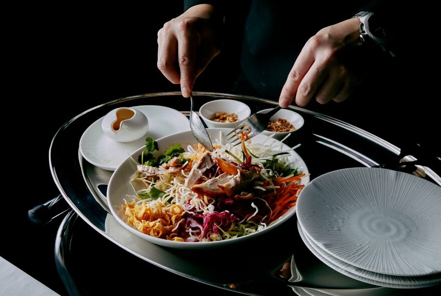 A silver platter with a share plate of asian salad, small side plates and condiment bowls, a pair of hands holding servingware.