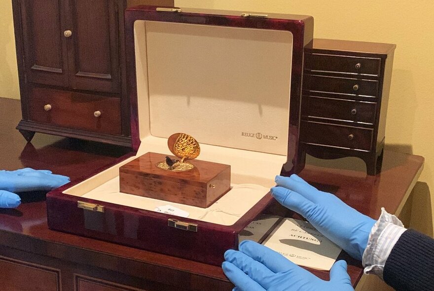 A polished wooden jewellery display case with an antique trinket inside, with three hands in blue rubber gloves carefully presenting it.