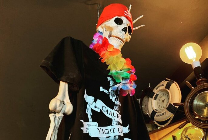Skeleton wearing a black t-shirt, red scarf on skull and floral necklace, with nautical decor on the wall behind the skeleton.