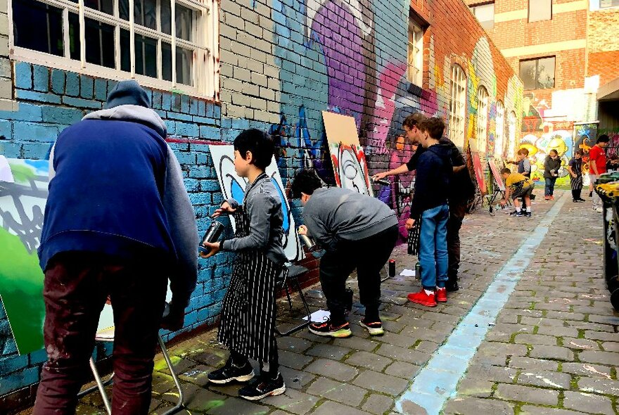 Row of children working on canvases along coloured brick wall.