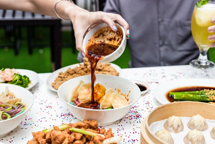 Someone pouring sauce over a bowl of dumplings on a table that contains lots of other dishes. 