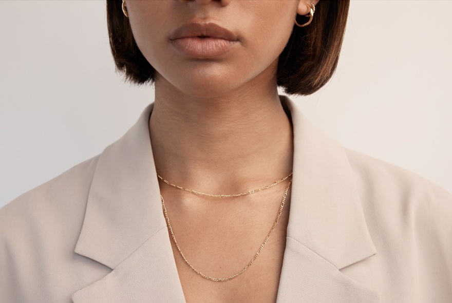 Close-up of a fine gold necklace and gold hoop earrings worn by a woman with short brown hair wearing a pale jacket.