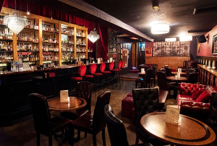 Interior of a speakeasy-style bar with rich wooden tones, red velvet bar stools and a deep red chesterfield surrounded by wooden circular tables and leather chairs.
