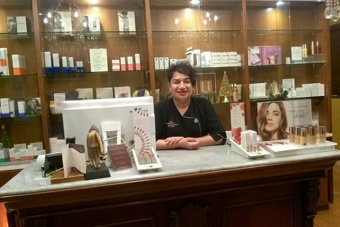 Smiling owner of beauty clinic leaning over the salon counter with glass shelves of beauty and skin products visible behind her.