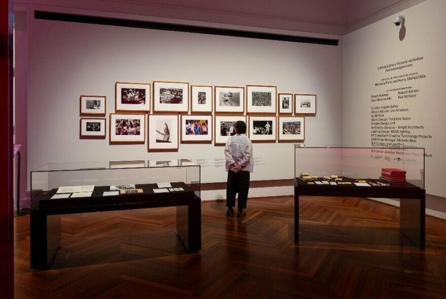 Installation view of the Rennie Ellis exhibition in a room in a gallery space, with a series of framed photographs displayed on a wall, with captions, and a person looking at them up close. 