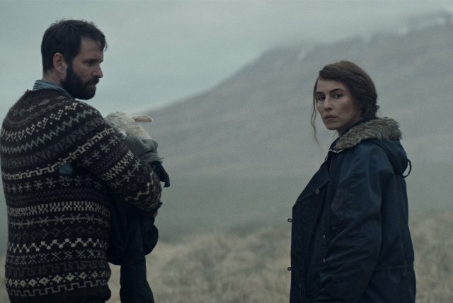 Man in a patterned knitted jumper holding in his arms a lamb wrapped in a blanket, standing next to a woman wearing a heavy overcoat, both of them in a bleak outdoor landscape.